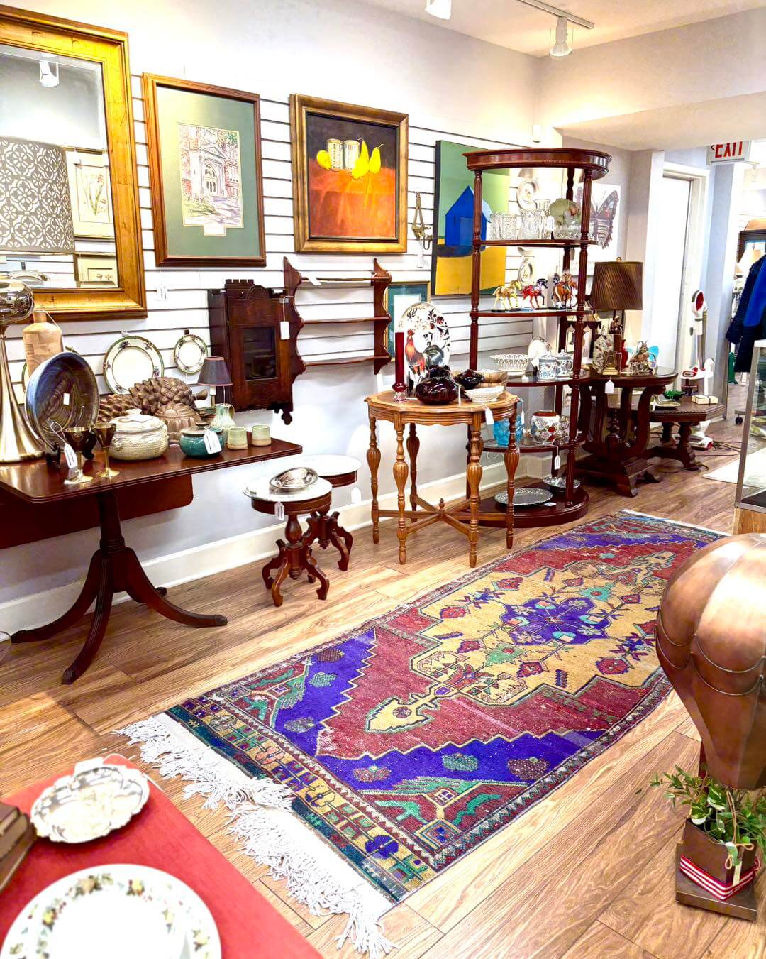 An antique shop interior inspired by a womans exchange, featuring wooden tables, decorative items, ceramics, framed art on the walls, and a colorful patterned rug on a hardwood floor.