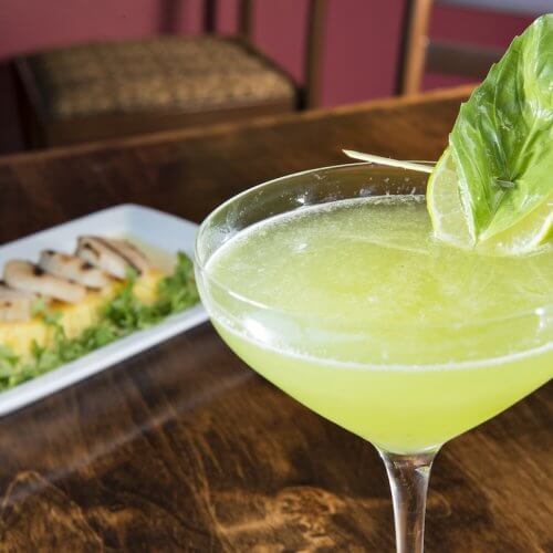 A basil martini garnished with a fresh basil leaf sits in a martini glass, while a plate of sliced grilled chicken and polenta rests in the background on a wooden table.