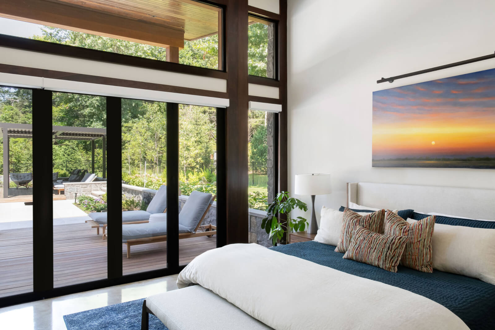 Modern bedroom by Beth Haley Design features large windows and sliding glass doors opening to a patio with lounge chairs. A landscape painting hangs above the bed, and greenery is visible outside.