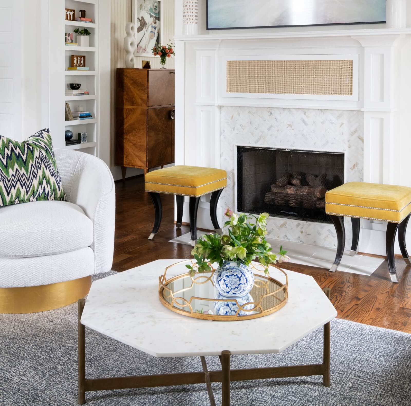 A living room designed by Beth Haley Design features a white armchair, a marble coffee table, and two yellow stools in front of a fireplace. A decorative vase with greenery adorns the table, while bookshelves create an inviting backdrop.