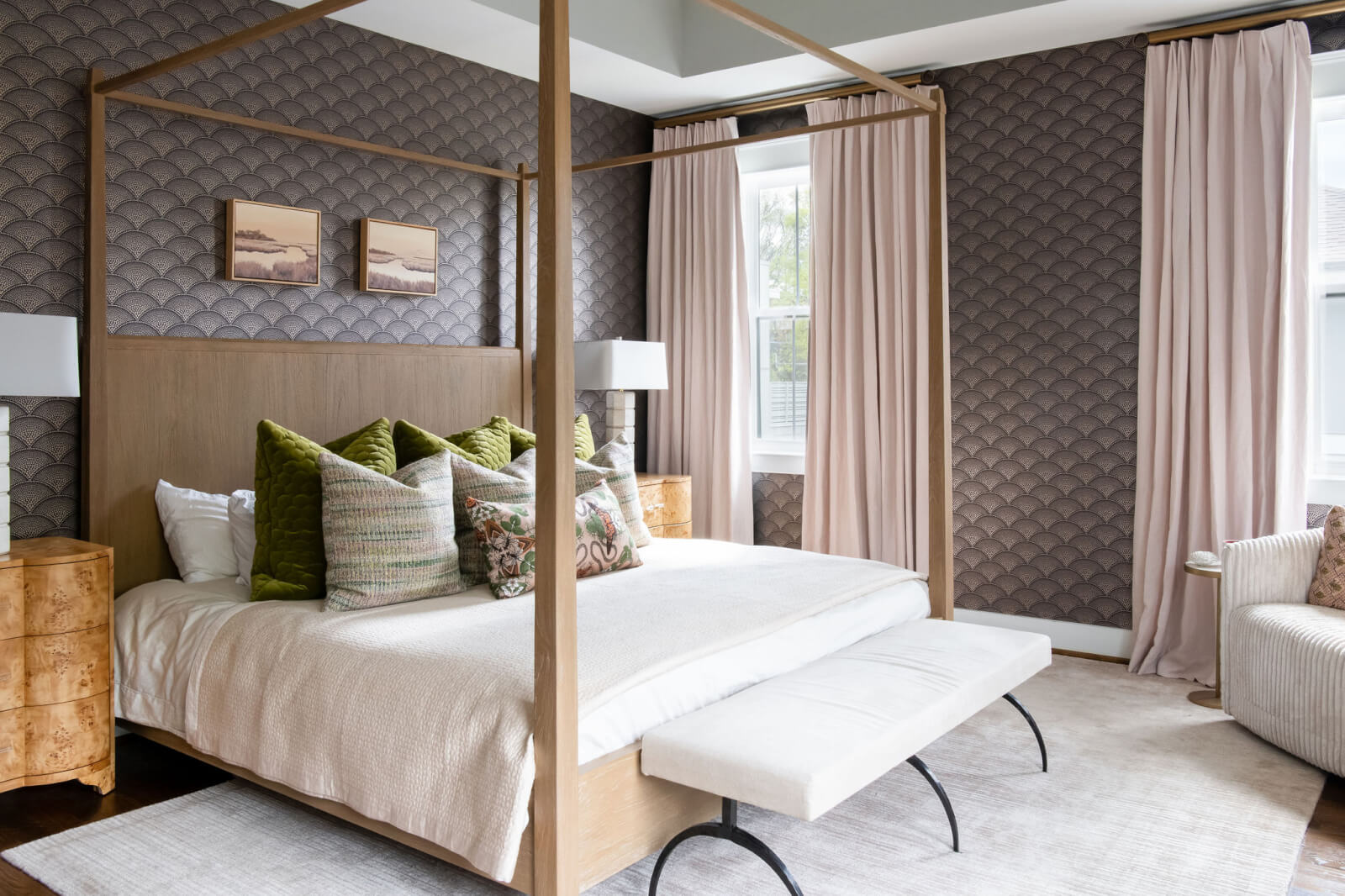 This charming bedroom by Beth Haley Design features a four-poster bed with patterned wallpaper and light curtains. Two small paintings grace the wall above the headboard, while a cushioned bench sits at the foot of the bed, complemented by elegant wooden nightstands.