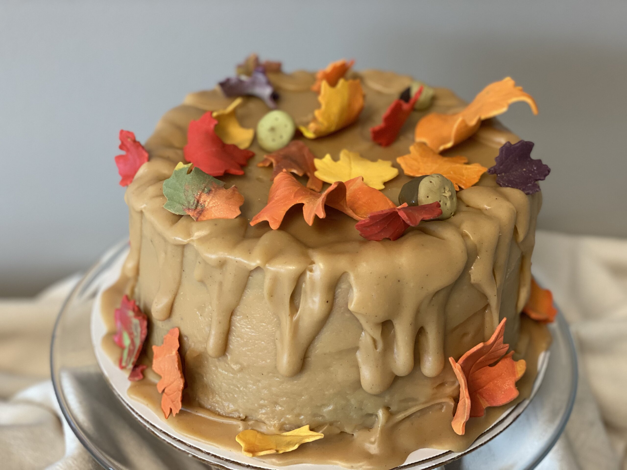 A round dessert design cake frosted with tan icing and a caramel drip, decorated with edible autumn leaves and small acorns.