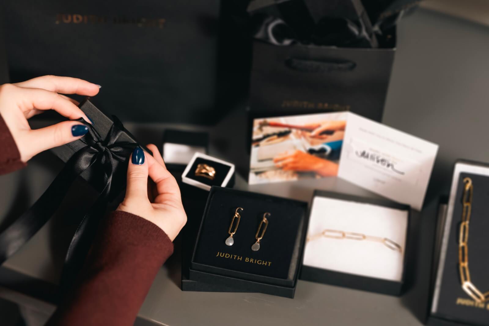 A person holds a black gift box surrounded by Judith Bright jewelry, with stunning earrings, bracelets, and elegant packaging scattered across the gray surface.