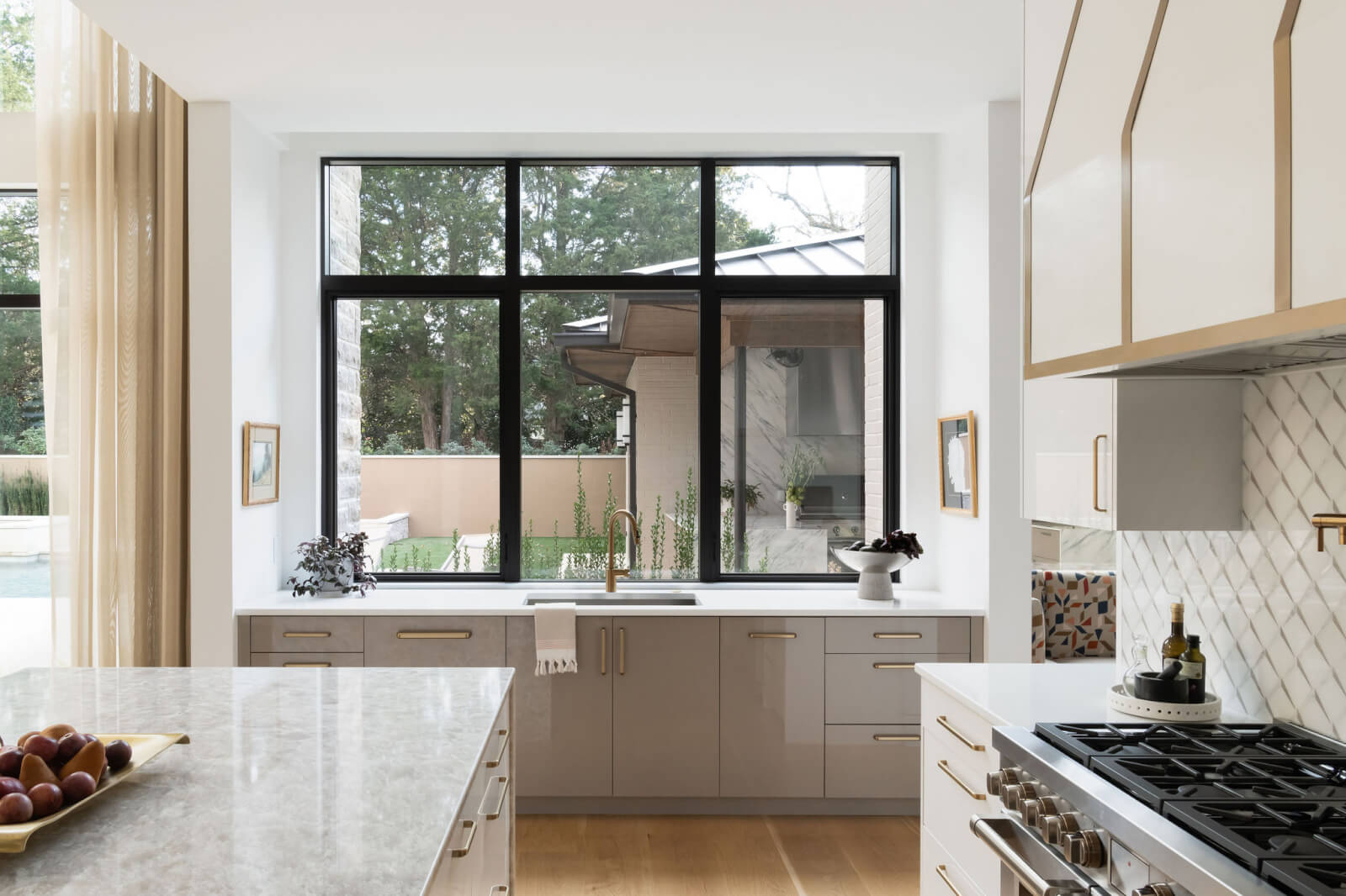 Modern kitchen designed by Beth Haley Design, showcasing marble countertops, a large black-framed window, and light cabinetry. The gas stove complements the setup beautifully. A sink is perfectly positioned under the window, flanked by potted plants and fruit bowls.