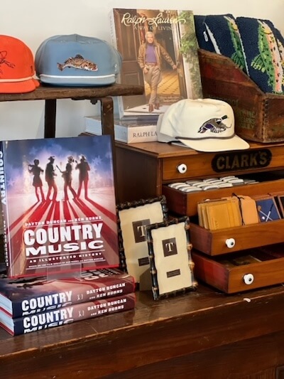 A display at White's Mercantile features books titled "Country Music," framed images, various hats, folded textiles, a Ralph Lauren book, all arranged on a wooden shelf alongside Clarks-branded items.