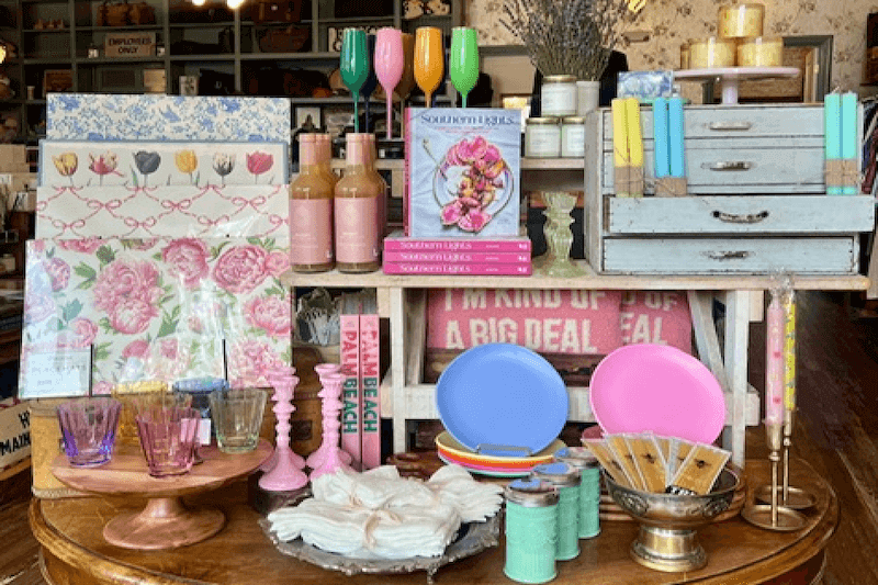 A display table at White's Mercantile features floral-themed books, decorative plates, colorful glasses, candles, and boxed matches in a retail setting. Pastel color tones dominate the arrangement.