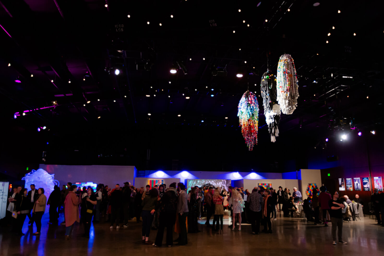 A crowd of people gathers in a large, dimly lit event space with colorful hanging decorations and OZ-themed artwork displayed on the walls.