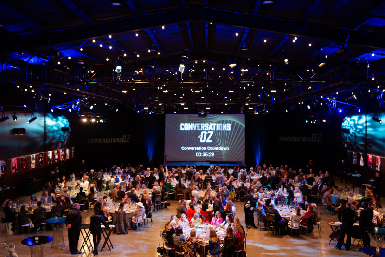 Large group of people seated at round tables in a dimly lit event hall, with an OZ-themed "Conversations OZ" countdown timer displayed on a screen at the front.