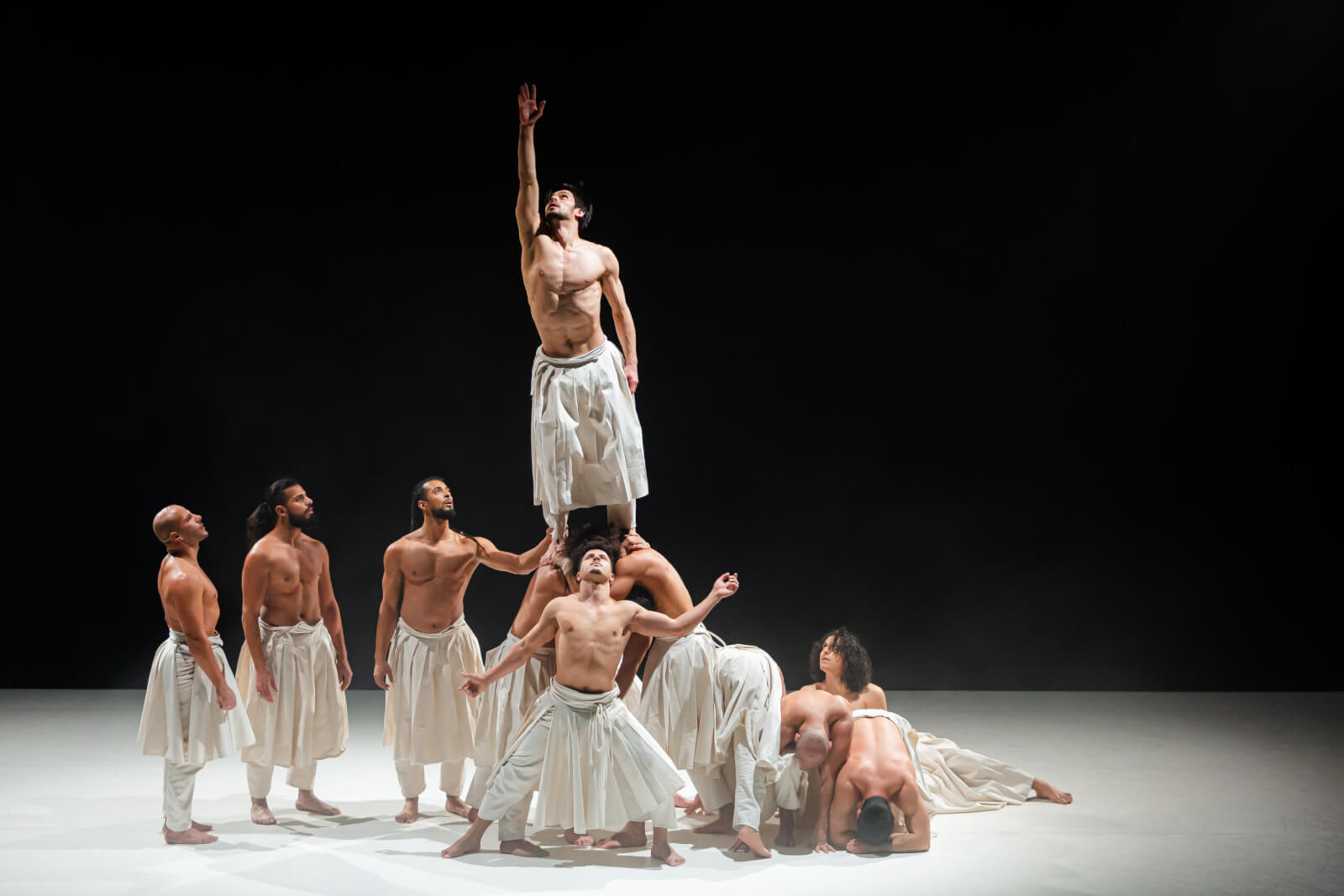 Eight barefoot dancers in white skirts perform on stage; one, elevated above the others, reaches upward with his right arm as the rest support or pose around him—evoking a dreamlike scene from OZ.