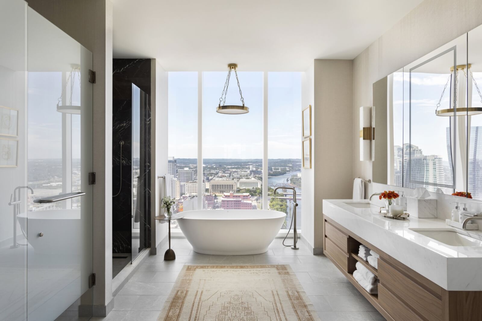 A modern bathroom designed with Providence Interiors flair features a freestanding bathtub by a large window, a glass shower to the left, and double sinks with a wooden cabinet on the right. The harmonious blend of style and function perfectly complements the stunning city view outside.