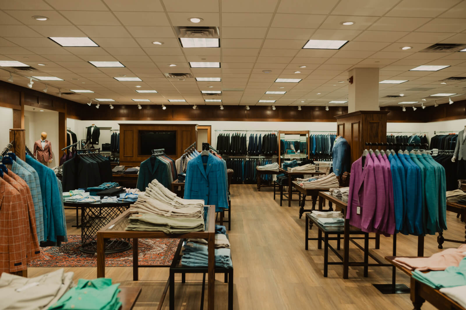 A men's clothing store interior at Oak Hall features suits, jackets, and trousers neatly displayed on racks and tables, all illuminated by bright overhead lighting.