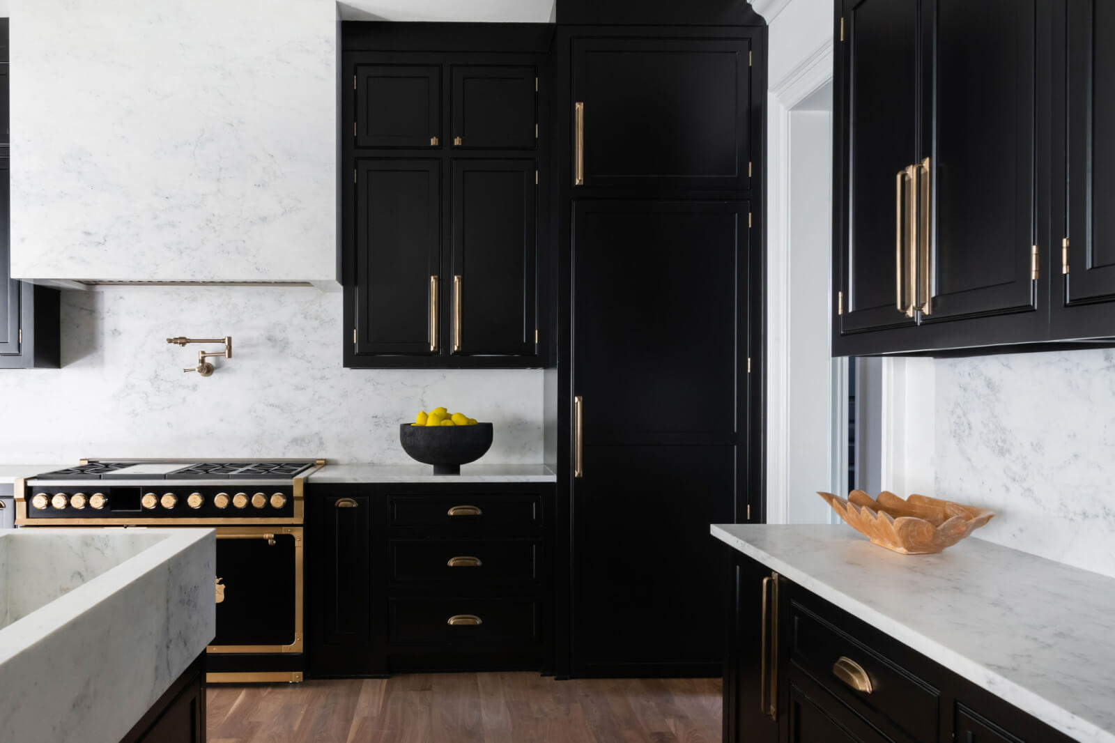 A modern kitchen by Sara Ray Interior Design features black cabinets, marble countertops, a gas stove with gold accents, and a bowl of lemons on the counter.
