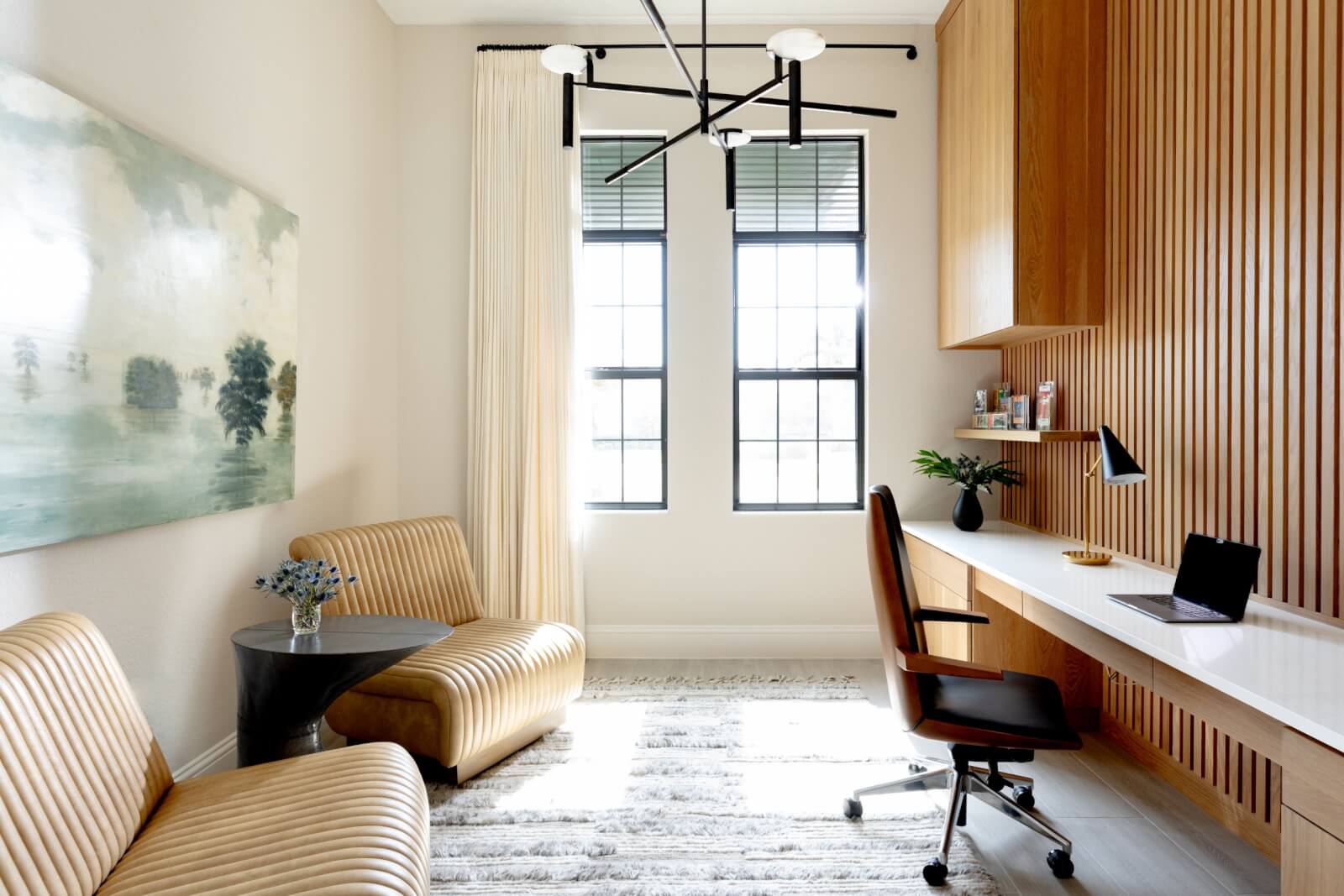 Modern home office with a minimalist design by Sara Ray Interior Design, featuring a desk, chair, built-in shelves, striped armchairs, a small table, large windows, abstract wall art, and a black chandelier.