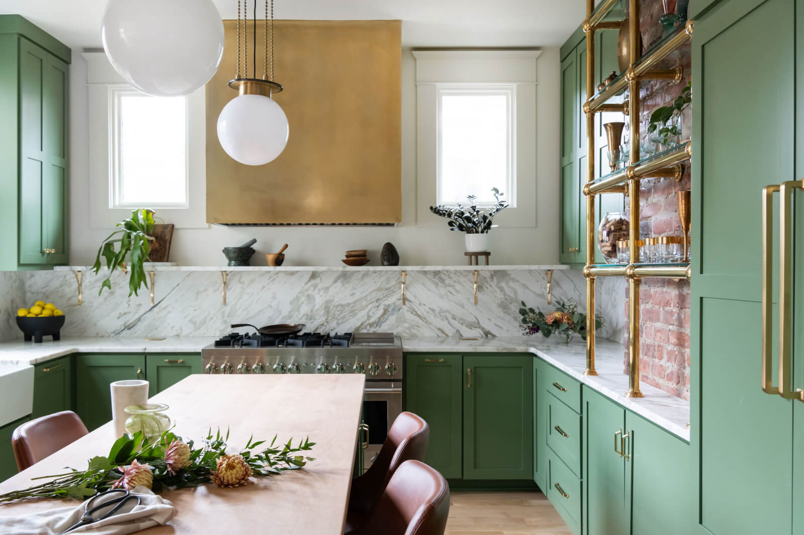 A modern kitchen by Sara Ray Interior Design features green cabinets, a marble backsplash, a gold range hood, and open shelving. A wooden dining table with a floral arrangement is centered in the room.