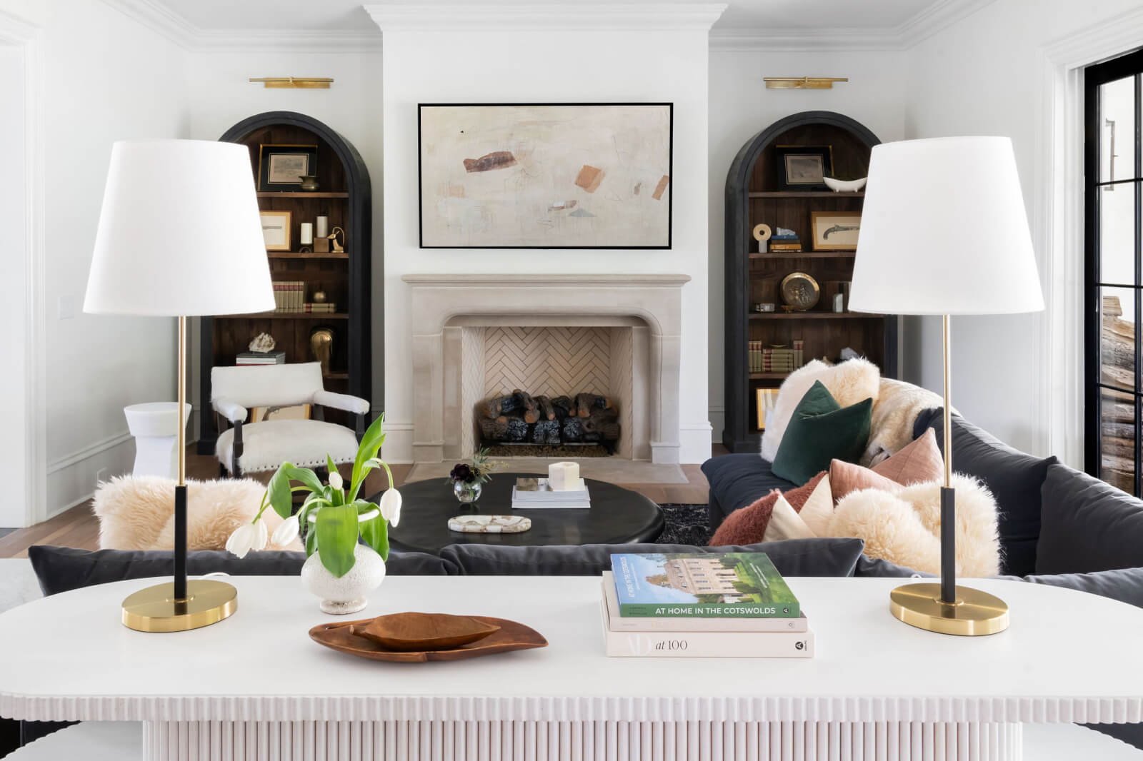 A modern living room designed by Sara Ray Interior Design features a central fireplace, two bookshelves, a dark sectional sofa with assorted pillows, a white coffee table, and a white table adorned with two gold-based lamps and displayed books.