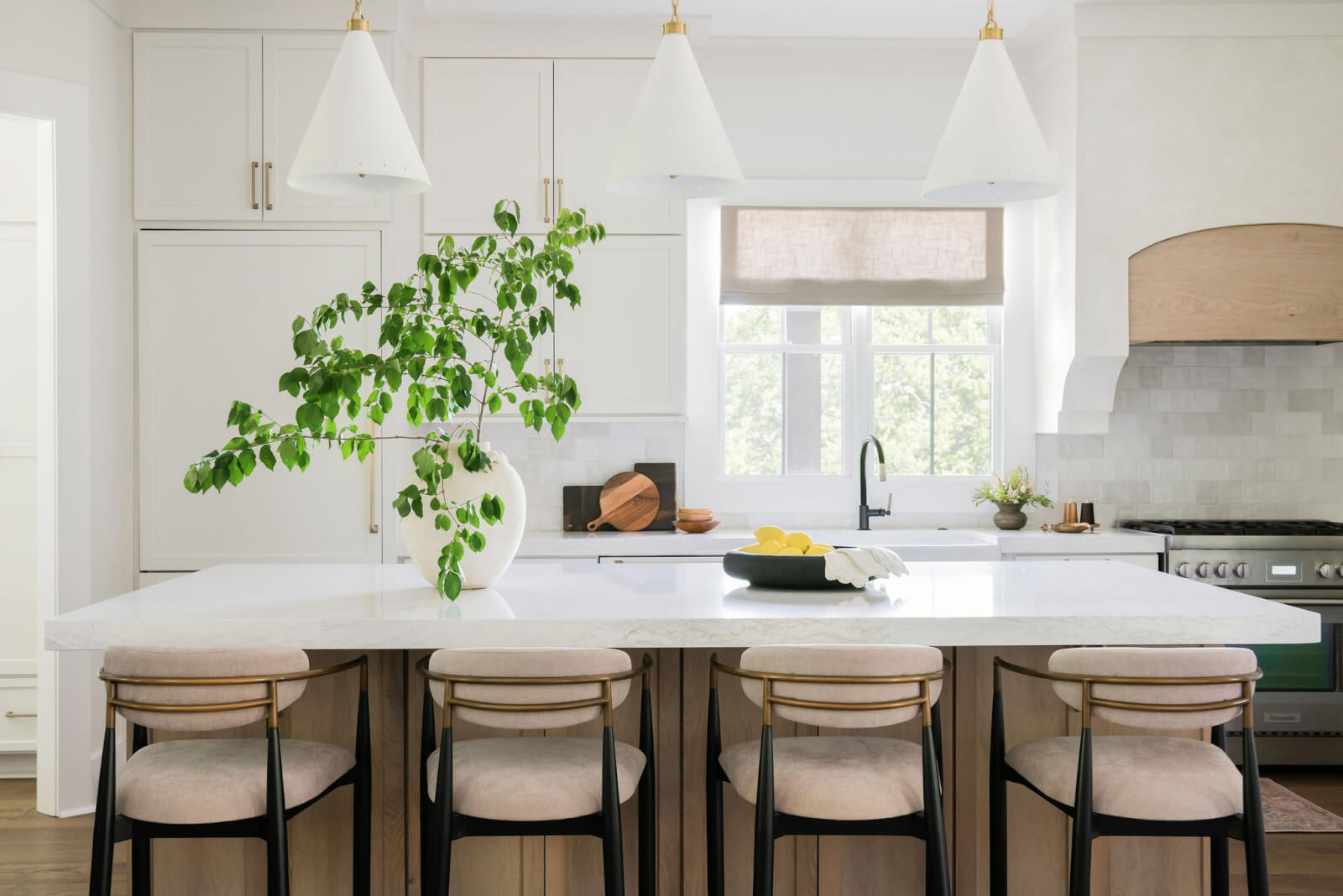Modern kitchen designed by Sara Ray Interior Design, featuring a large white island, four bar stools, pendant lights, a vase with green branches, a bowl of lemons, and pristine white cabinetry. A window with a beige shade graces the space above the sink.