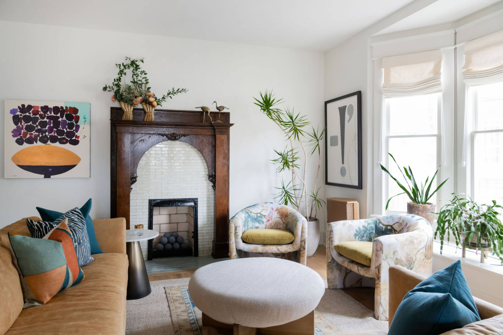 A cozy living room by Sara Ray Interior Design featuring a beige sofa with colorful cushions, two patterned armchairs, a round ottoman, and a decorative fireplace with a mirror above it. Plants and modern artwork adorn the space.