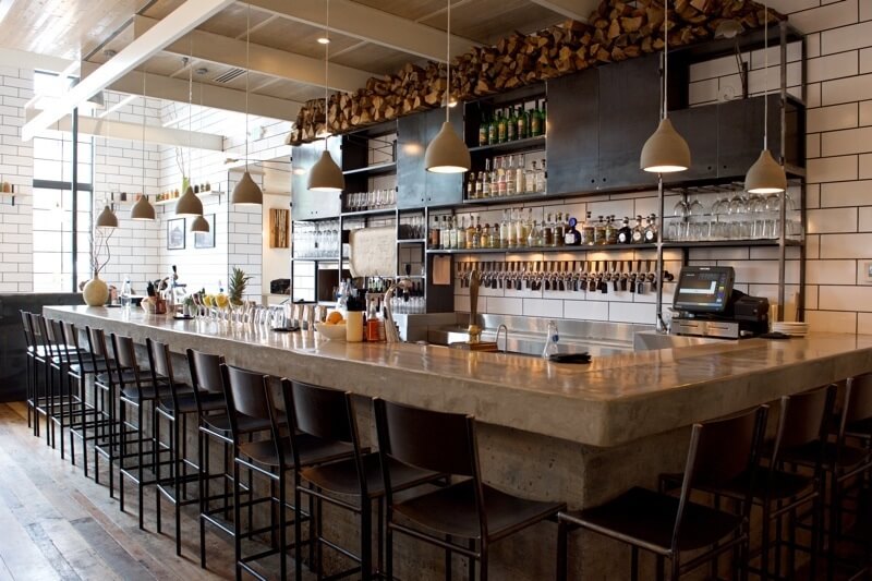 Metal plays a leading role as log holders, table bases, aerial rods for suspended globe lights, banquettes and booths, chairs, bar stools and shelving behind the bar, which is poured concrete, as are the lights over the bar.