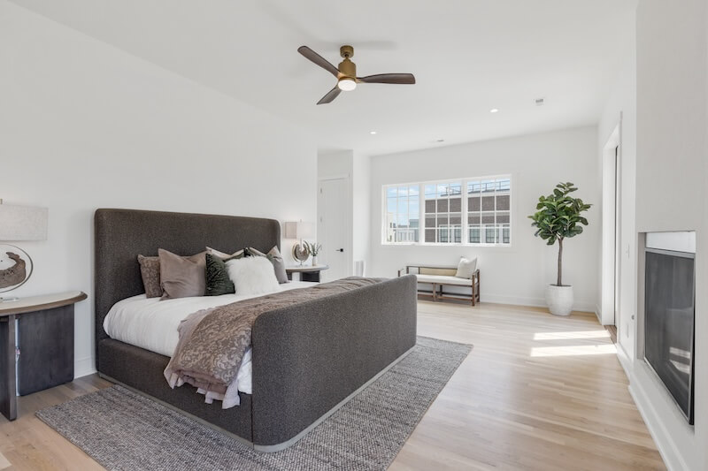 Modern bedroom featuring a large bed with a dark headboard, white walls, a fireplace, and expansive windows, accented by a potted plant and a sitting area.