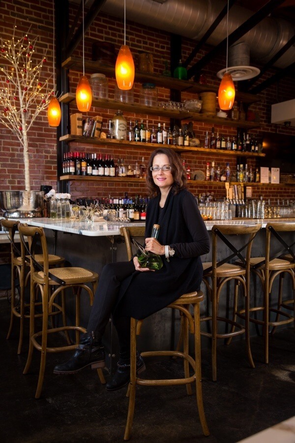 Idie sits at the beautiful, bohemian bar at OvenBird restaurant with a porron, a traditional glass wine pitcher native to Catalonia and other regions of Spain. Enjoy a porron with friends at OvenBird!