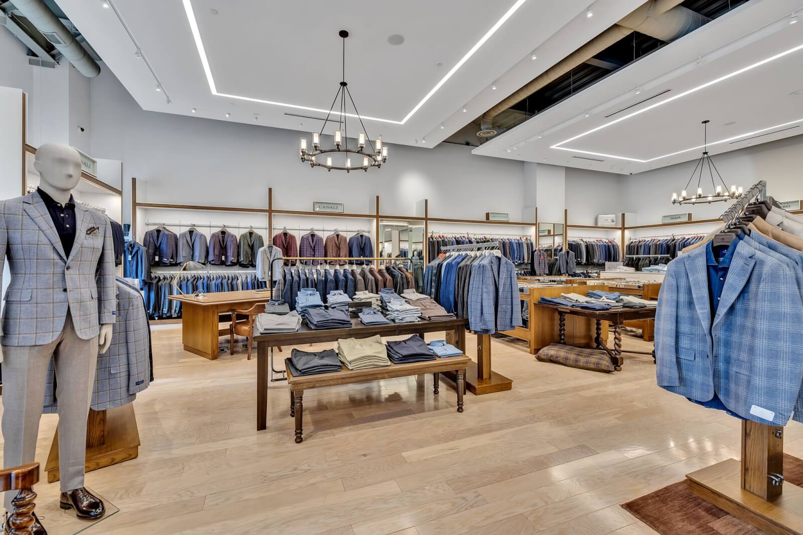 Men's clothing store interior at Oak Hall with neatly arranged suits, shirts, and pants on racks and tables, mannequin in a checkered suit, and bright overhead lighting.