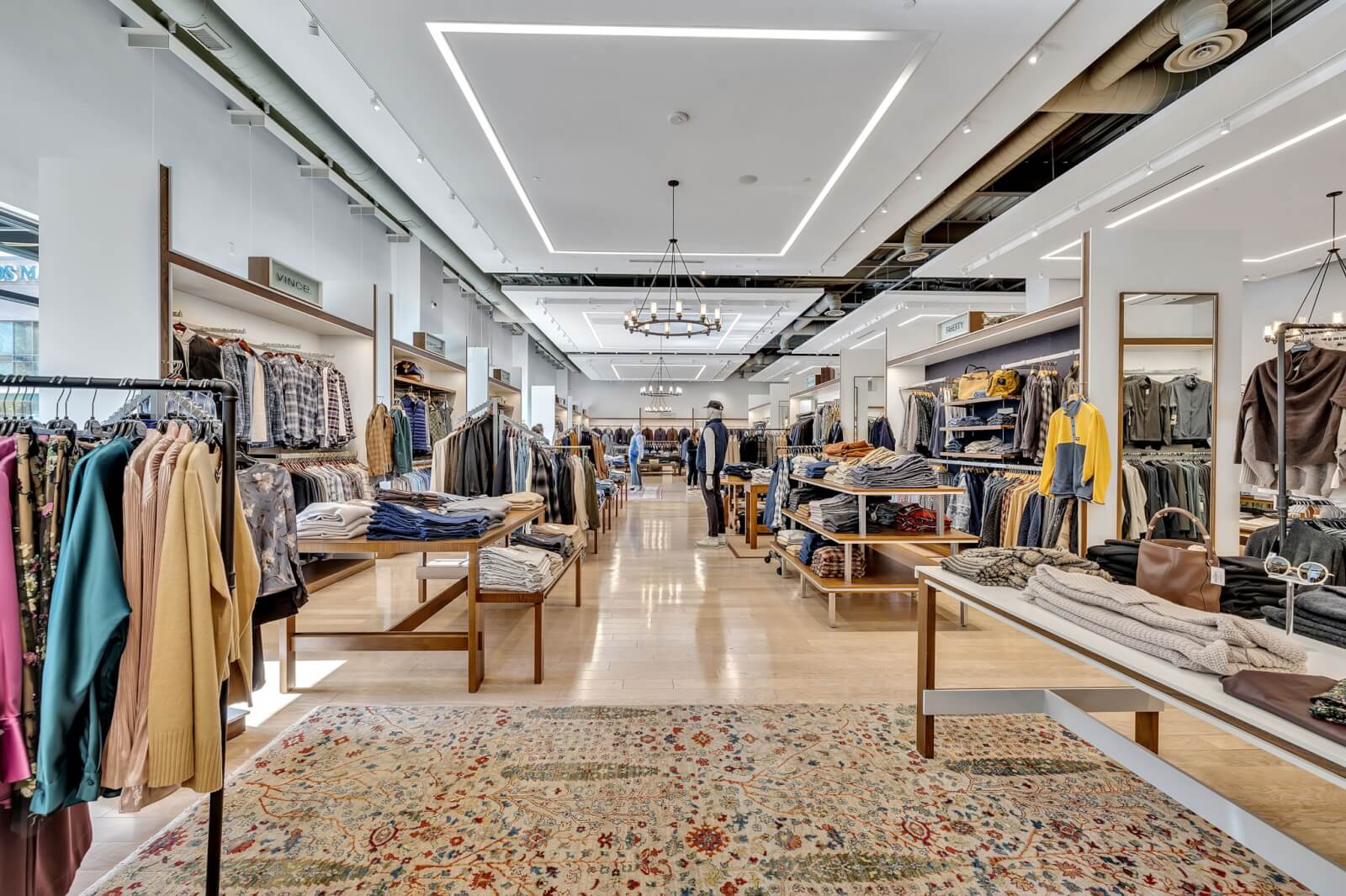 A modern clothing store interior at Oak Hall with neatly arranged racks and shelves of clothes, bright lighting, large mirrors, and a patterned rug on the wooden floor.