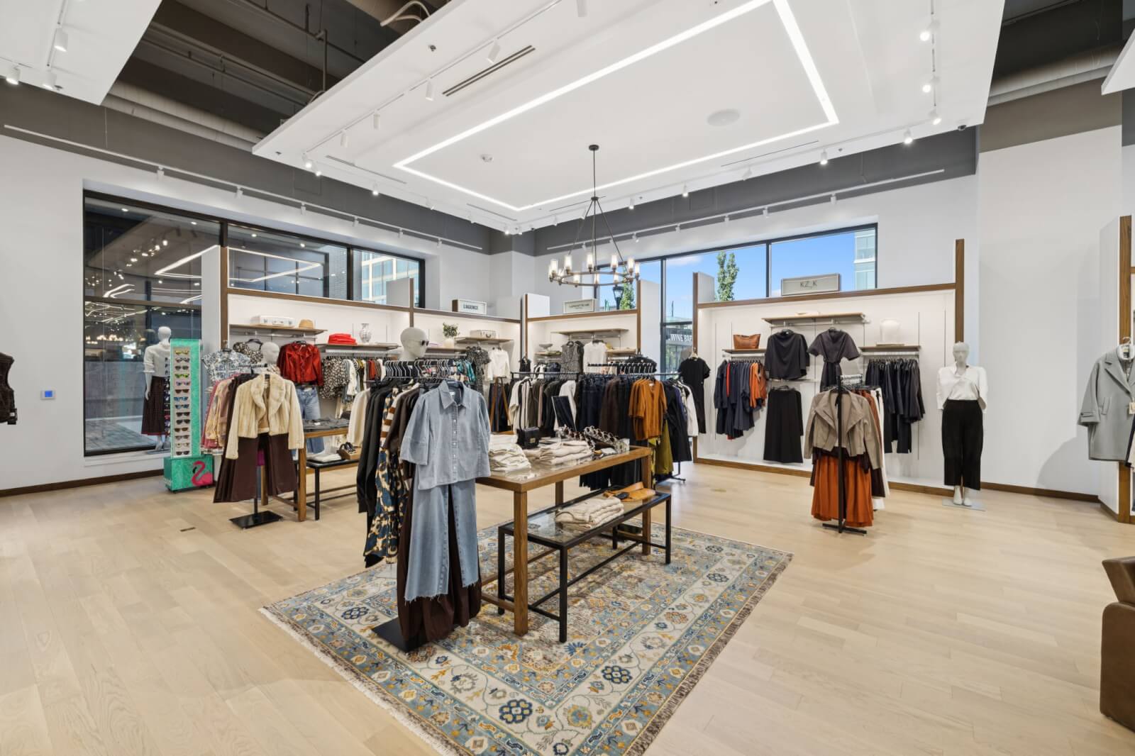 Modern clothing boutique, reminiscent of Oak Hall, with mannequins and neatly arranged racks of women's apparel displayed on light wood floors and a decorative area rug under soft ceiling lights.