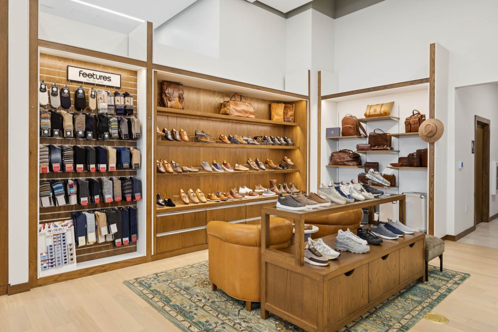 An Oak Hall shoe and accessories store display with shelves of shoes, socks, and leather bags, a wooden bench with sneakers, and a leather armchair on a patterned rug.