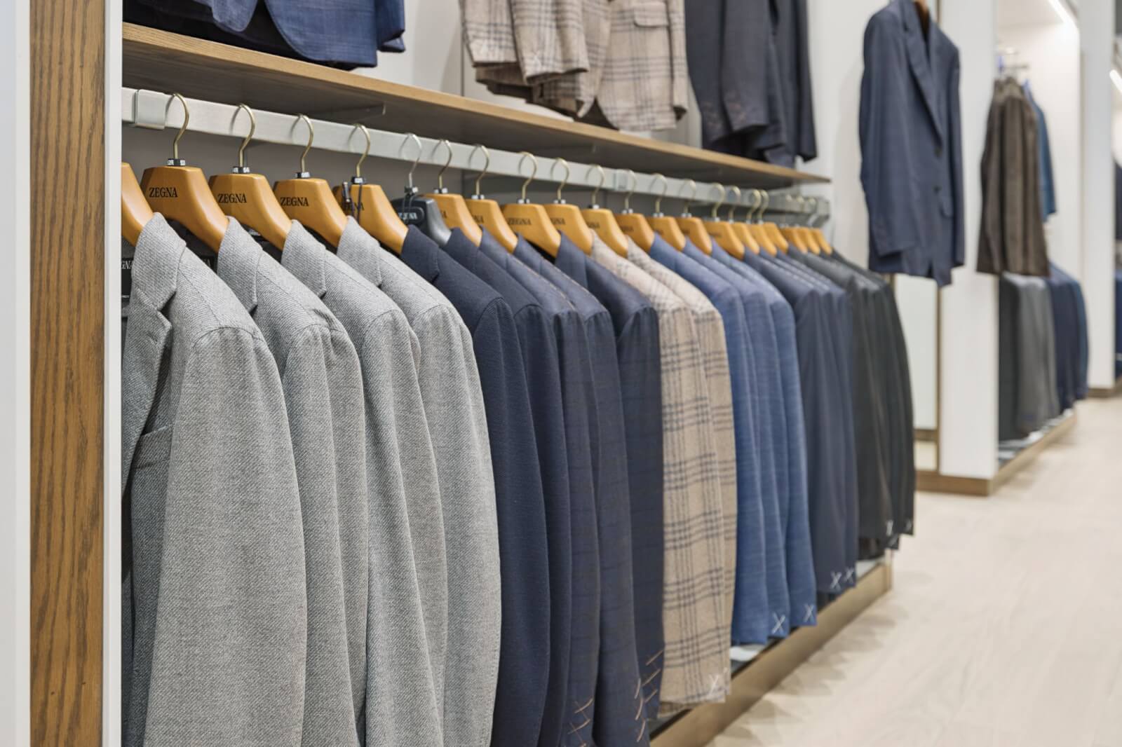 A row of men's suits in various shades of grey, blue, and plaid hangs neatly on wooden hangers at Oak Hall, showcasing timeless style in a refined clothing store setting.