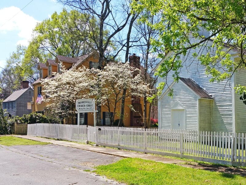 The Mooresville Church of Christ, circa 1854, stands beside historic homes in vibrant yellows and blues.