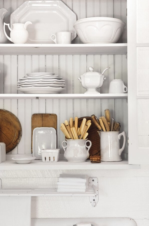 This cream colored over-the-sink cupboard displays white serving ware and bone-handled cutlery in plain sight and within easy reach. The utilitarian display with a casual mix of whites offers a relaxed, effortless charm.