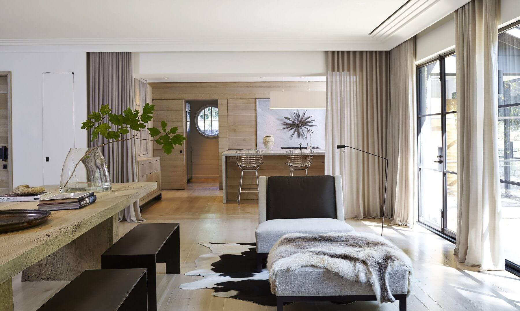 This kitchen and dining room open floor plan features a custom dining table in antique white oak by Christian Liaigre chairs and ottoman. The kitchen includes Bertoia counter stools, a striking suspension lamp by Christian Liaigre and a Brutalist wall sculpture. Image: Jean Allsopp