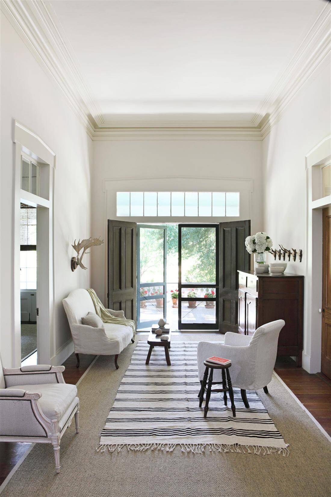 The side entry features lovely, old-school screen doors leading onto the covered porch. Inside, a striped kelim anchors traditional elements, such as the antique Louis XV settee, mid-century Louis XVI-Style Bergère, the antique French buffet and primitive antique stools. Image: William Abranowicz 
