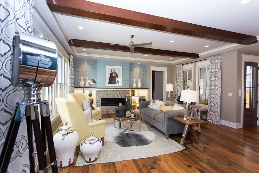 Design elements of this great room include chrome and natural wood, as seen in both the side table and round coffee table as well as lamps throughout the room. High-backed, pale yellow chairs evoke traditional simplicity and the low-backed gray, chenille covered sofa adds a comfortable, inviting touch. Custom, batik-influenced drapes and a cowhide rug add texture and the pale-blue accent wall creates an eye-catching backdrop for a contemporary print.