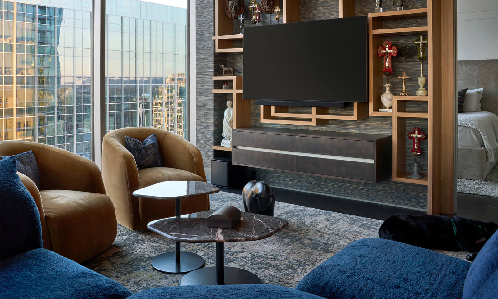Modern living room by interior designer Marcelle Guilbeau, featuring blue sofas, gold chairs, marble coffee tables, wall-mounted TV, decorative shelves, and large windows with a cityscape view. Bedroom partially visible on the right.