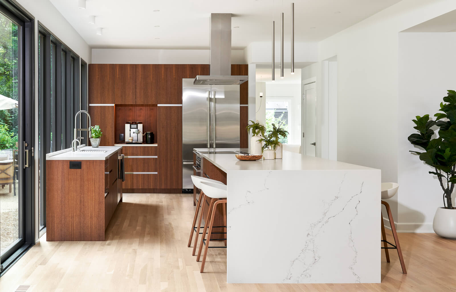 Modern kitchen with wood cabinetry, a marble island with bar stools, stainless steel appliances, and large glass doors allowing natural light—an inviting space that showcases refined interior design.