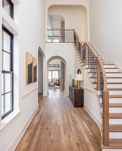 A bright, spacious hallway designed by Marcelle Guilbeau Interior Design features light wood flooring, a staircase with a wooden handrail, arched doorways, wall art, and a console table with a lamp, leading to a living area with large windows.