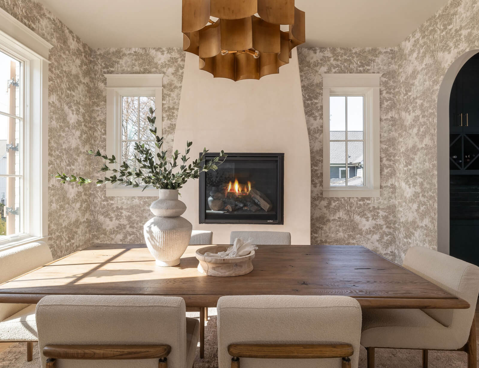 A dining room by interior designer Marcelle Guilbeau features a wooden table, six upholstered chairs, a vase with green branches, a fireplace, two windows, and a large wooden ceiling light fixture.