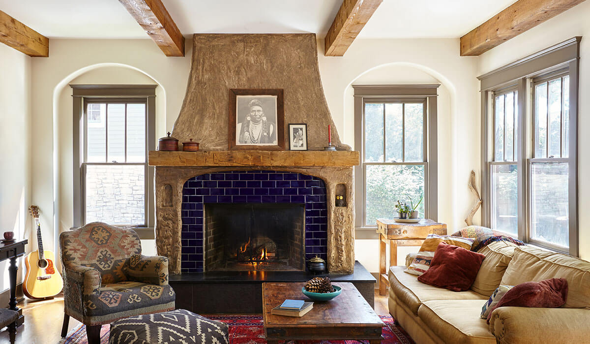 Cozy living room with exposed wood beams, a stone fireplace with blue tiles, a large mantel, two armchairs, a beige sofa, and natural light—classic touches by interior designer Marcelle Guilbeau.