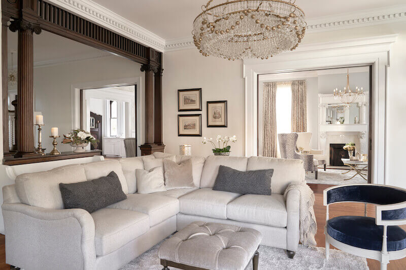 A well-lit living room, expertly designed by Marcelle Guilbeau, features a beige sectional sofa with gray pillows, a tufted ottoman, a large chandelier, framed artworks on the wall, and views into a dining area adorned with another chandelier.