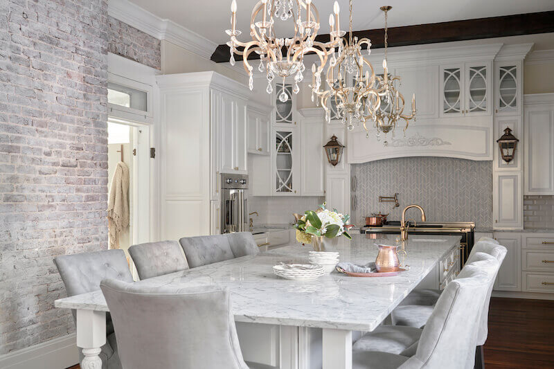 A bright kitchen designed by Marcelle Guilbeau features a large marble island with grey upholstered chairs, a chandelier, white cabinets, stainless steel appliances, and a brick accent wall.