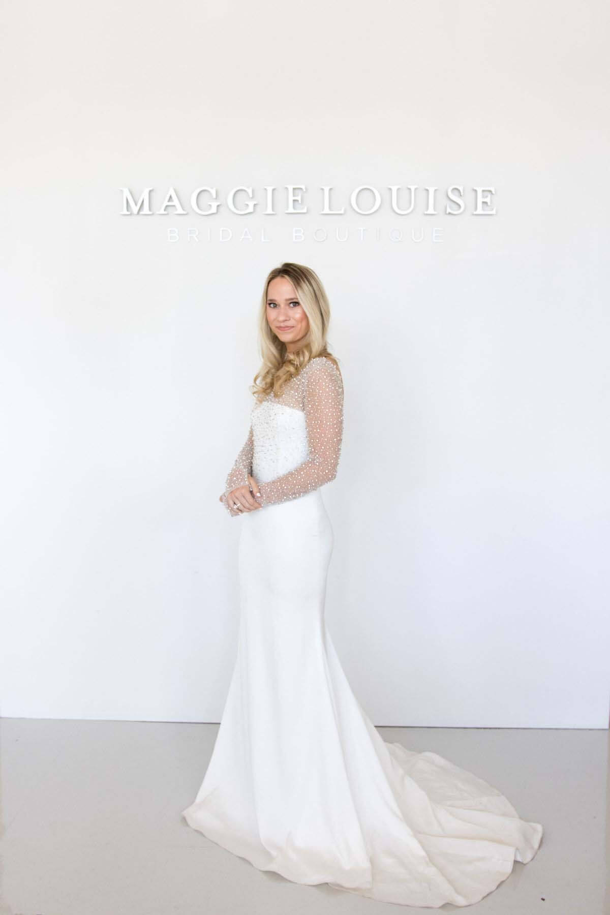 Maggie Louise has been focused on bringing in crepe styles, both full crepe dresses as well as a combination of crepe and detailing. Image: Maggie Louise Bridal Boutique Bride modeling Maggie Louise wedding dress in front of store's sign