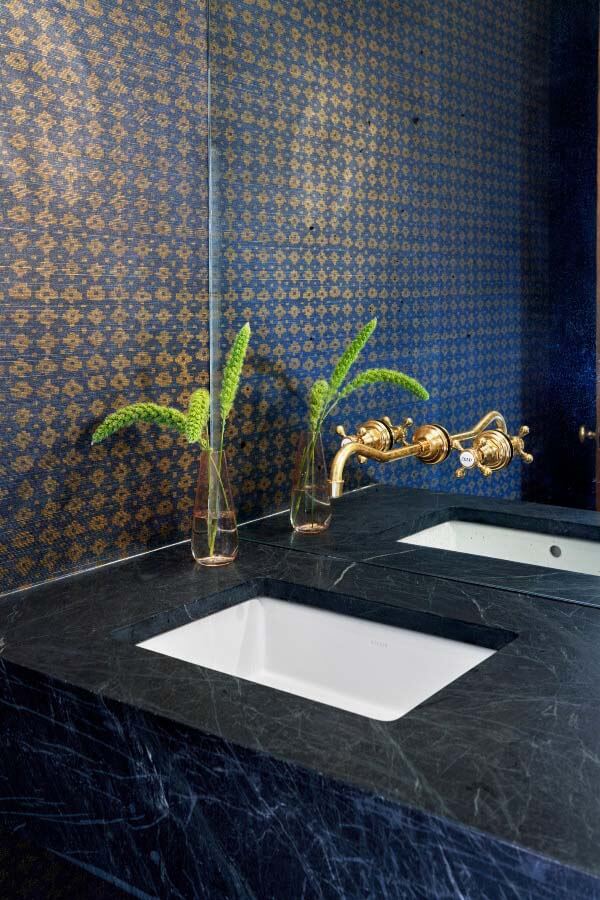 Somewhat atypical for the house, this powder room is a bit more on the dark and moody side. The Phillip Jeffries grasscloth adorning the walls adds elegance to the windowless space, and a custom-built floating soapstone vanity goes from wall to wall. Local vendor, Blue Door, created the antiqued mirrored wall behind the vanity. Powder room with navy and gold wallpaper, gold fixtures