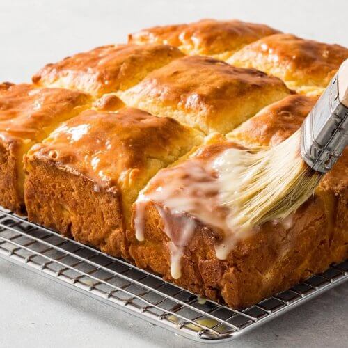 A baked pull-apart bread, reminiscent of buttery biscuits, rests on a cooling rack as it is being brushed with a glaze.