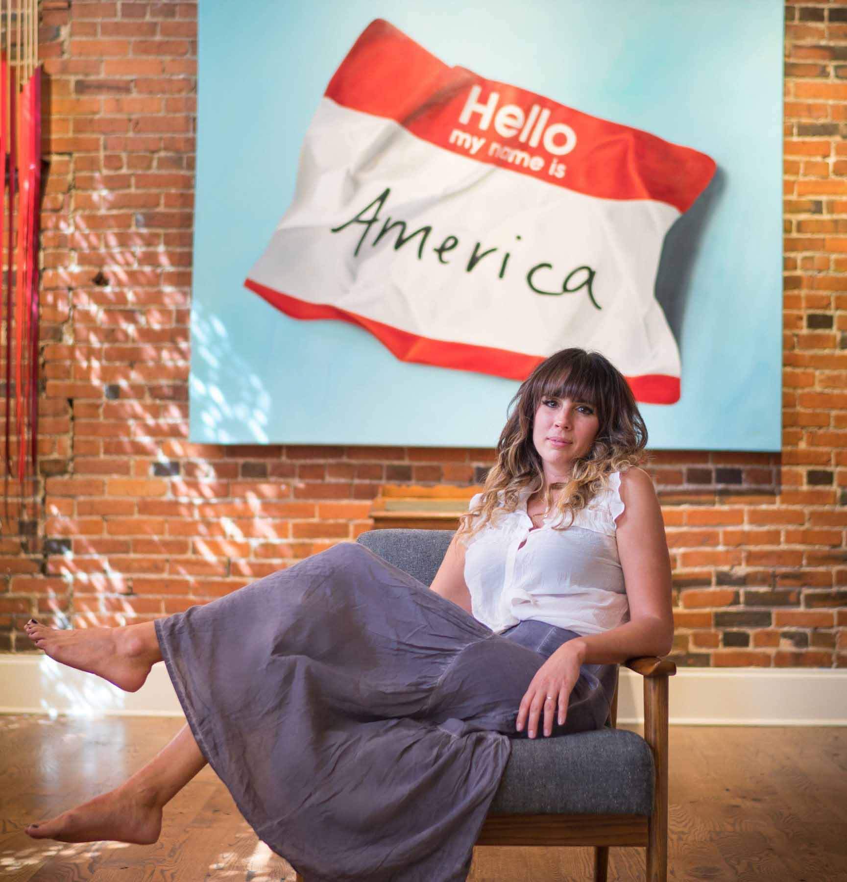 Artist Kristin Llamas in front of artwork saying "Hello my name is America"