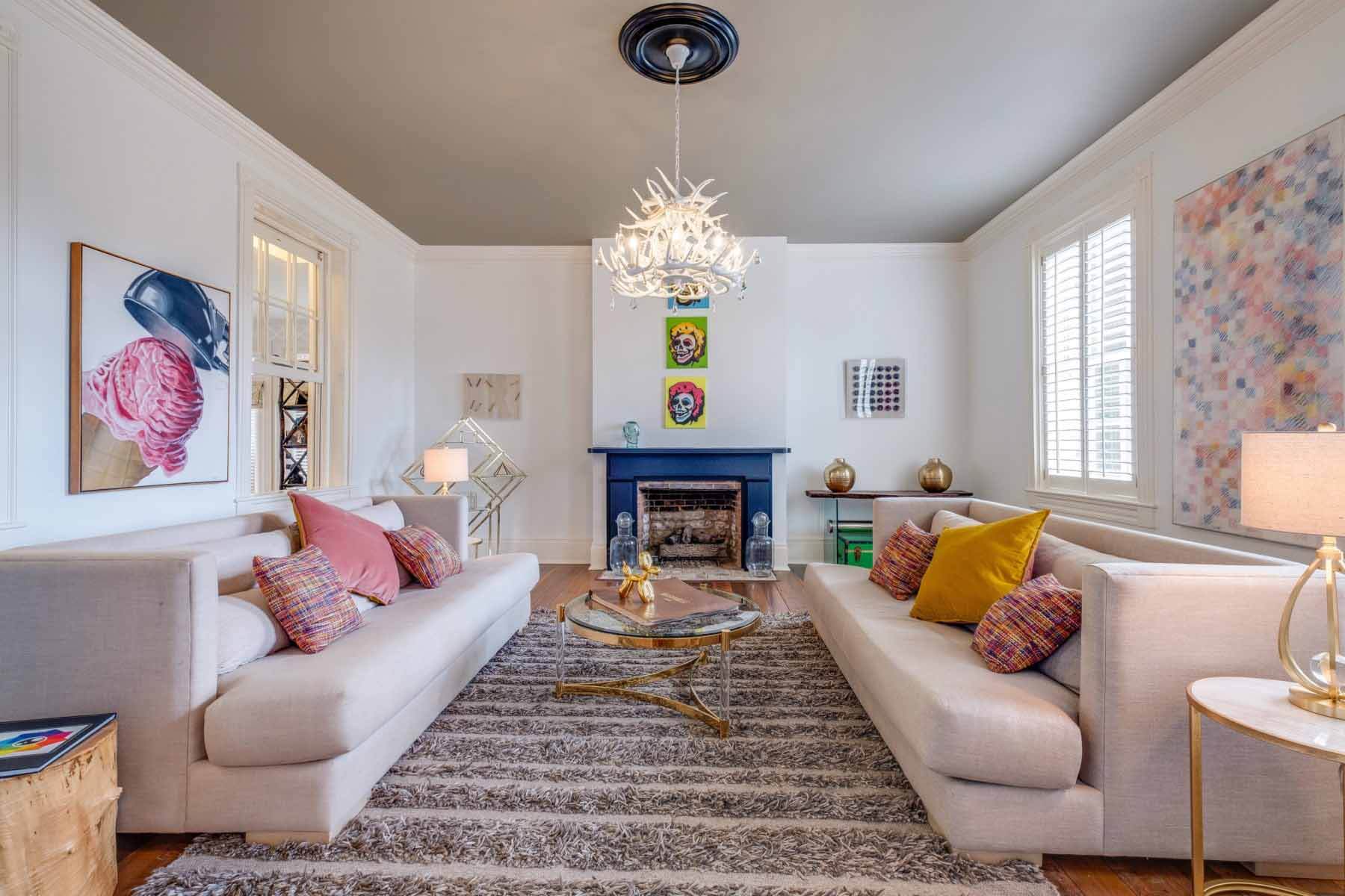 With a pop of color from the royal blue fireplace, this sitting room, formerly the women's parlor, is chock full of vibrant street art and interesting curiosities such as a book of local maps from 1986. "The female parlor has a shorter fireplace than the male's parlor," Therese says, " and there are a bunch of black divots on the floor near the fireplace, where somebody dropped the coal." Image: Zach Harrison Photography Seating area with white couches, royal blue fireplace mantel and antler chandelier