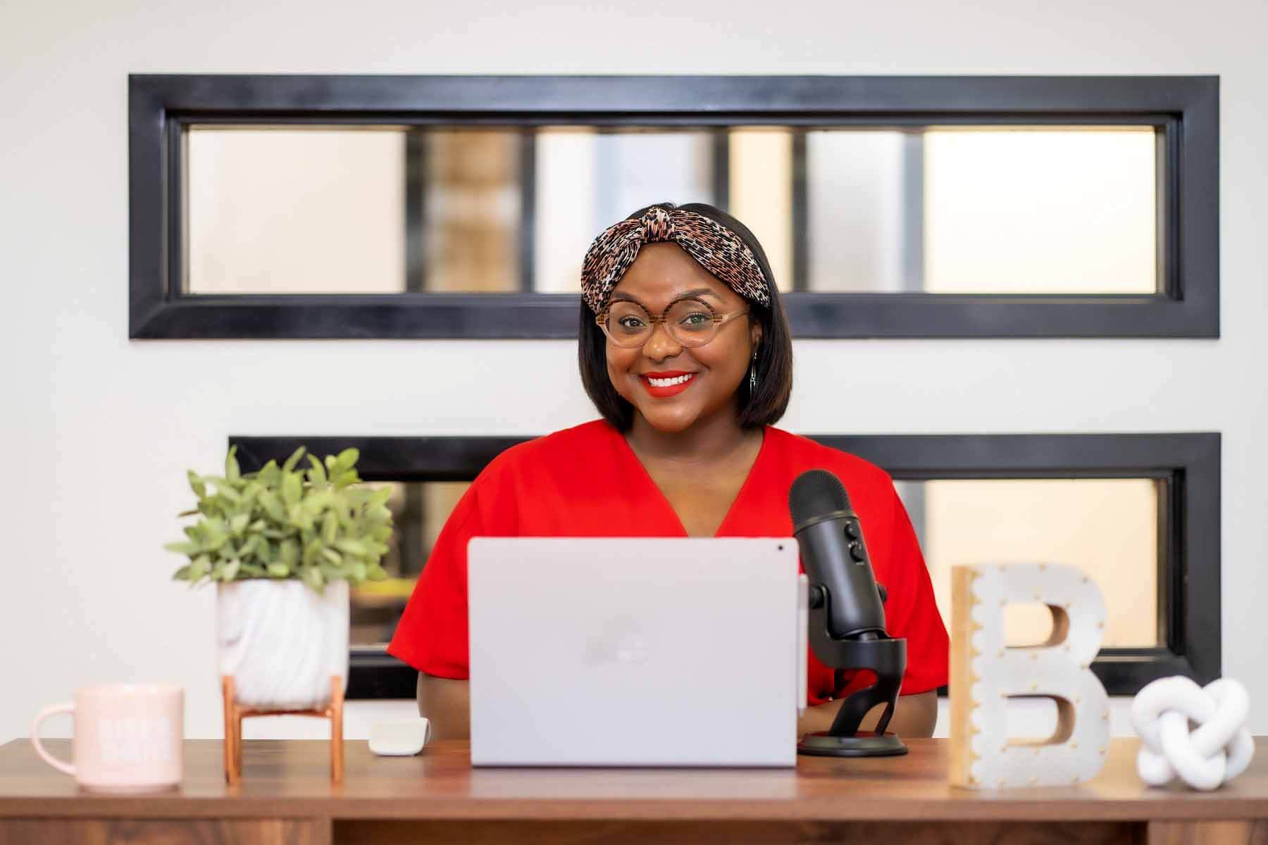 Brittany Cole in a red blouse, sitting at her desk with her computer and a microphone