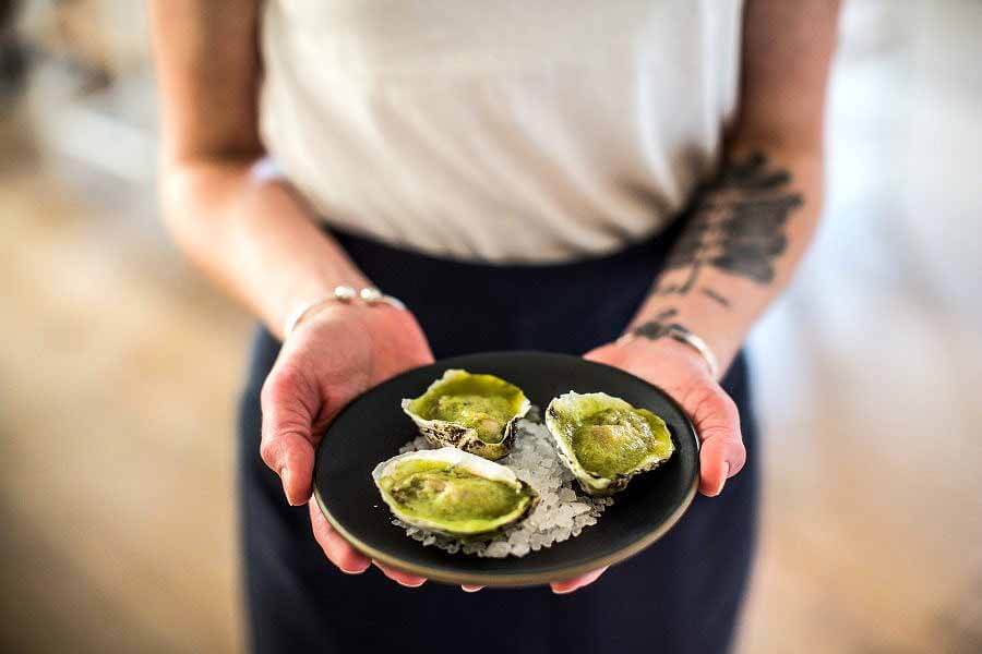 20 Epic Spots to Grab Dinner at the Bar in Nashville - 6 A woman holding a plate of three oysters on salt, coated in a green sauce.