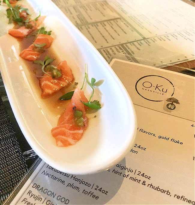 A long white plate featuring thinly sliced salmon with sauce and microgreens from O-ku