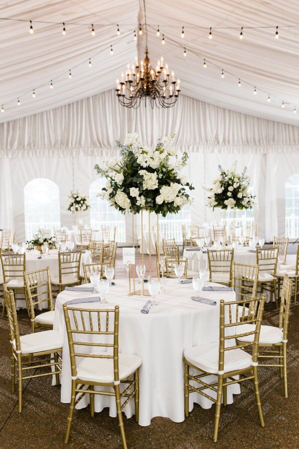 Wedding tent decorated with chandelier, round tables, gold chairs and white florals.